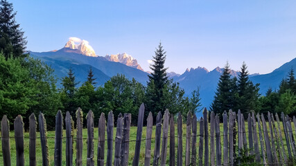Fototapeta premium A wooden fence in front of a lush pasture with a view on the first sunbeams reaching the peaks of Ushba in Caucasus, Georgia. Cloudless sky above the high and snow-capped mountains. Daybreak.