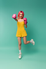 full length of young woman with pink dyed hair in boxing gloves jumping and screaming on blue