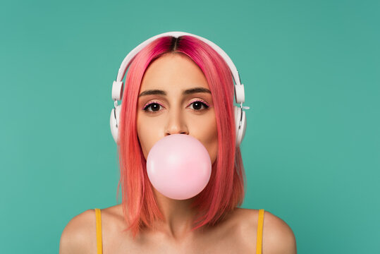 young woman with pink dyed hair in wireless headphones blowing bubble gum isolated on blue