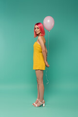 full length of smiling young woman with pink dyed hair holding balloon on blue