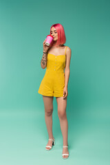 full length of young woman with pink dyed hair holding paper cup while drinking coffee to go on blue