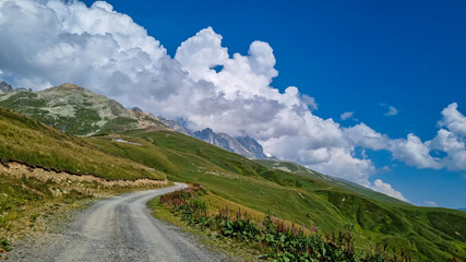 Naklejka premium A gravelled road in high Caucasus mountains in Georgia. The road leads to a ski resort on a steep slope. Thick clouds in the back. Lush pastures on the sides. Barren peaks.