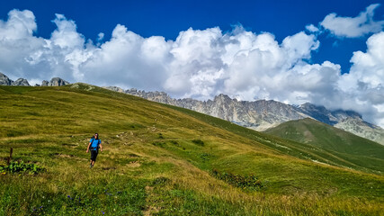 Fototapeta na wymiar A man hiking along a narrow pathway in high Caucasus mountains in Georgia. There are high glaciers in the back. Thick clouds above the sharp peaks. Lush pastures on the sides. Barren peaks.
