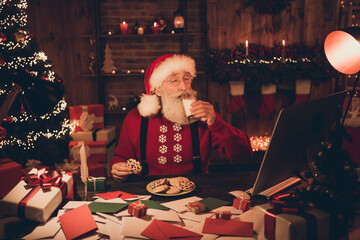 Photo of aged santa claus happy positive smile drink milk eat cookies workshop eve time midnight atmosphere magic indoors