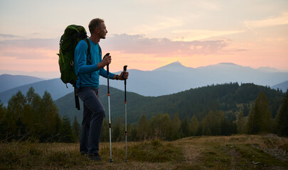 Man traveler against backdrop of mountain hills and pink cloudy evening sky at sunset. Hiker with trekking poles and touristic backpack standing on glade and admiring mountain fairytale landscape.