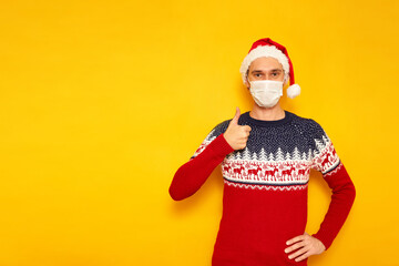 Fototapeta na wymiar man in Christmas sweater with reindeer, medical mask, New Year's hat, a Santa Claus hat, gives a thumbs up, likes, OK sign. isolated yellow background with space for text. concept holiday, gifts, sale