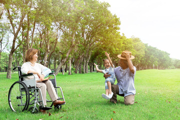 Asian family enjoy relaxing in the park, Grandmother sitting on the wheelchair and Grandfather playing together with little cut girl in the park, Green nature background.