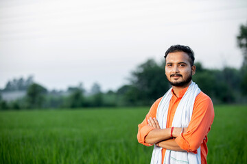 Happy Indian young man farmer giving side pose standing in field with hands crossed wearing kurta, smiling village male peasant looking on camera in farm, greenery, blur background, copy space.