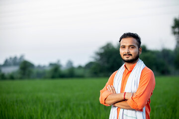 Happy Indian young man farmer giving side pose standing in field with hands crossed wearing kurta,...