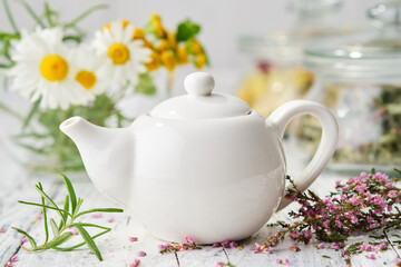 White tea kettle of healthy herbal tea, daisy and heather flowers on white wooden table. Alternative herbal medicine.