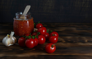Tomato sauce made of tomatoes, garlic and horseradish in a transparent jar with a wooden spoon. Cherry tomatoes, garlic head and basil leaves on dark wooden boards