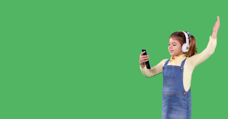 Portrait of young little school girl standing against green background holding the smart phone in her hand and listening the music using headphones, raised her other hand up.