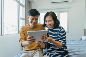 Happy elderly Asian couple smiling and looking at the same tablet on the bed, at home concept - retired man and woman using technology - lockdown and quarantine lifestyle