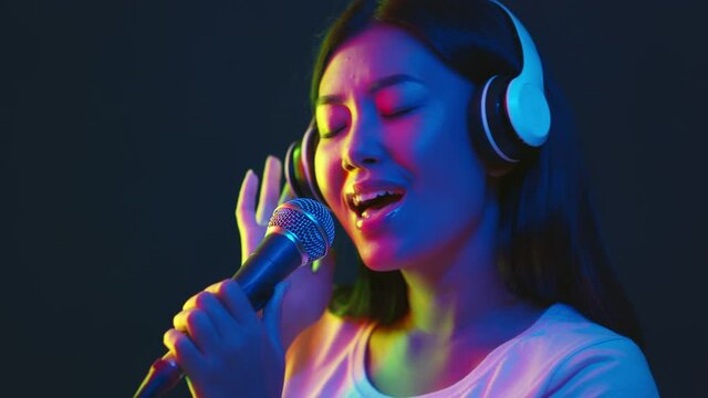 Carefree Singer. Young asian lady in wireless headphones singing favorite song at microphone in bright neon lights