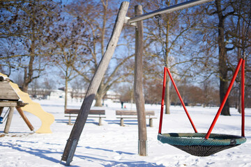 Large swing on a playground in the park, trees and building in the background. red dominates. Close up. Winter with snow in germany.
