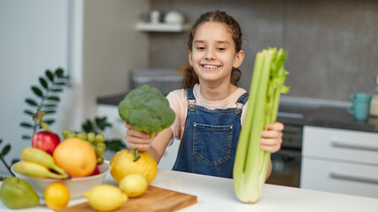 Obraz premium Smiling pretty little girl stand near table in the kitchen, holding green broccoli and celery in hands.
