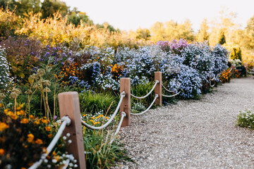 Pebble path next to a fence made of wood and rope in a garden full of various flowers. 