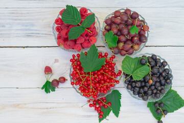 Different garden berries in plates on a white wooden background. Ripe juicy berries of red currant, black currant, gooseberry, raspberry with green leaves