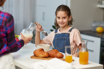 The happy little girl looking at camera having healthy breakfast together with mother in the kitchen.