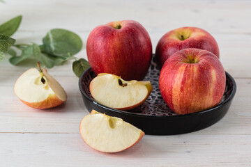 In a round black dish on white wooden table are three juicy beautiful ripe red apples of the early winter variety Gala. Next to the slices of the cut fruit