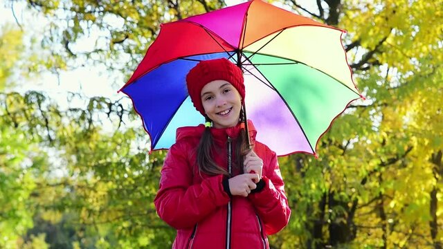 A smiling girl in a red hat and a red jacket walks with a rainbow umbrella in nature. yellow leaves in the background. Autumn. Atmosphere