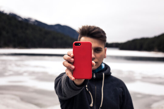 Young man holding red cell phone at a lake in winter