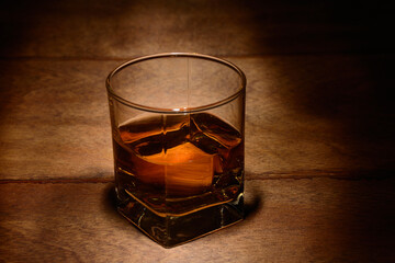 A glass of whiskey on a wooden table in a dark room lit by a weak light