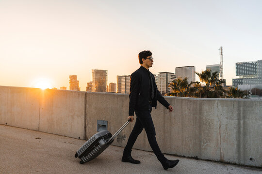 Spain, Barcelona, young businessman with rolling suitcase at sunset