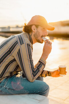 Tattooed man with coffee to go eating  doughnut at sunset