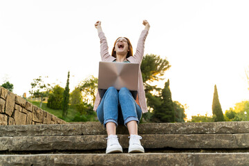 Young redheaded woman using laptop and cheering, sitting on steps in a park
