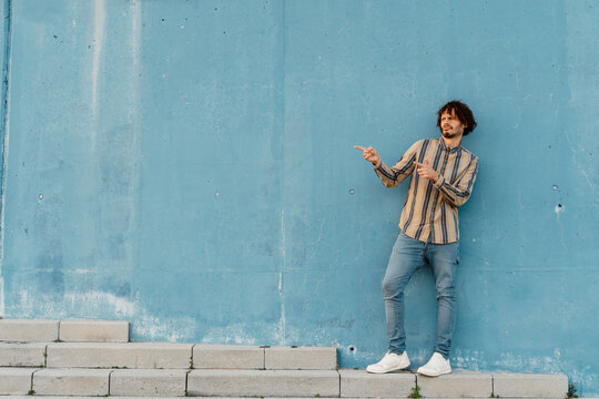 Bearded man wearing striped shirt standing ibn front of light blue wall gesticulating