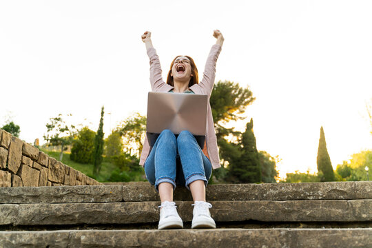 Young redheaded woman using laptop and cheering, sitting on steps in a park