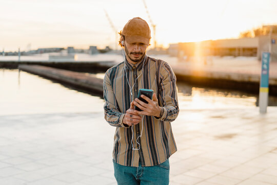 Man wearing baseball cap listening music with earphones and smartphone at sunset