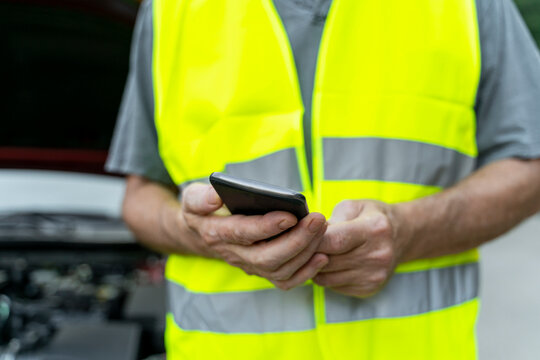 Close up of a senior man standing at his broken car wearing a safety vest and using his smartphone