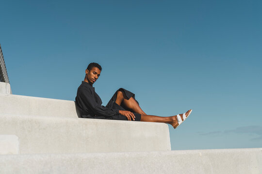 Young man wearing black kaftan sitting on concrete stairs under blue sky