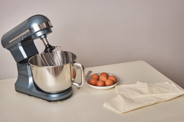 Electric mixer on the table with a metal bowl, next to brown eggs in a bowl and fabric, towel. Against the background of a gray-white wall, in the kitchen. The concept of healthy, natural, homemade