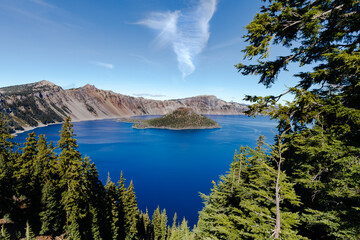Scenic wide landscape of deep blue water of Crater Lake, Oregon, with lush green trees and bright blue sky and wispy white clouds.