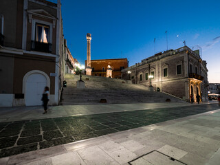 Illuminated Roman column on steps amidst buildings in Brindisi against sky at night