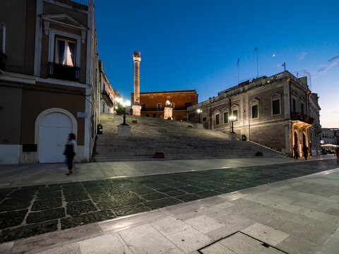 Illuminated Roman column on steps amidst buildings in Brindisi against sky at night