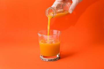 Woman's hand pours orange juice from a bottle into a glass, orange background