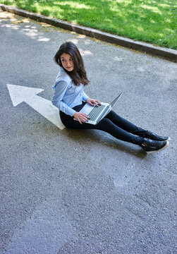 Young woman with laptop sitting on arrow sign on street