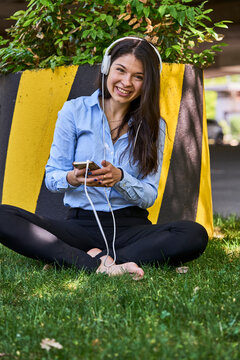 Portrait of smiling woman sitting barefoot on a meadow listening music with headphones and cell phone