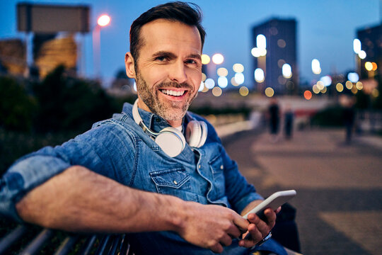 Portrait of a smiling man using his smartphone while sitting on a bench in the evening