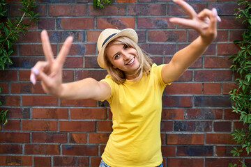 Portrait of happy young woman standing at a brick wall making peace sign