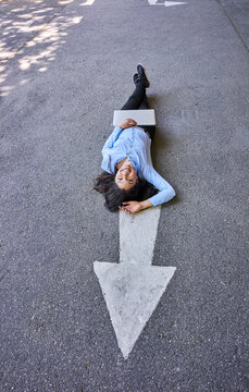 Laughing young businesswoman with laptop lying on arrow sign on street