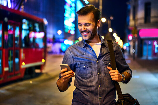 Smiling man with wireless headphones using smartphone while walking through the city at night