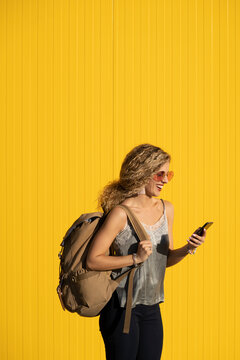 Laughing young woman with  backpack looking at cell phone in front of yellow background