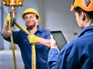 Industrial worker fixing hoist sling on indoor crane, female colleague controlling with digital tablet