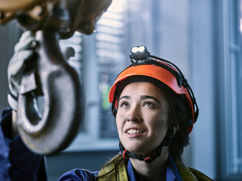Young female industrial worker with safety helmet taking hook of indoor crane