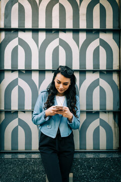 Portrait of young woman with long black hair standing in front of patterned wall using smartphone